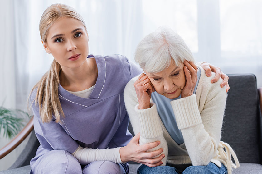 Young nurse looking at camera while embracing senior woman sitting bowed