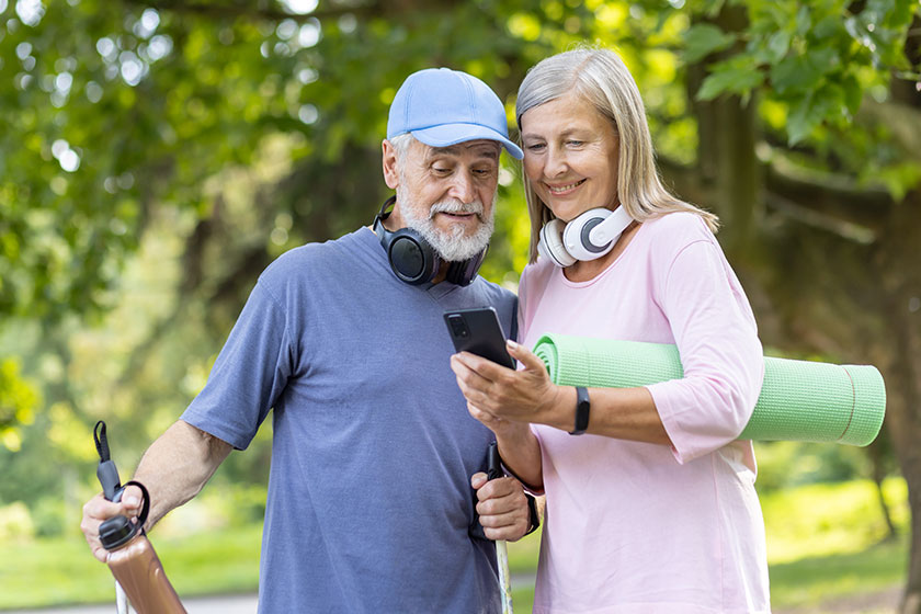 Happy mature man and woman in sportswear with yoga mat and headphones looking at a phone in a sunny park setting