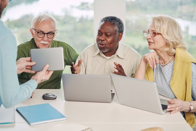 A small group of multicultural senior students in a classroom are looking at tablet with explanation and using laptops